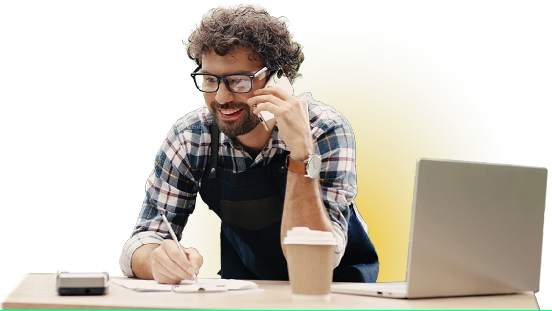 Mężczyzna oparty o biurko zapisuje notatki. Na biurku leży kubek z kawą, komputer oraz terminal płatniczy.